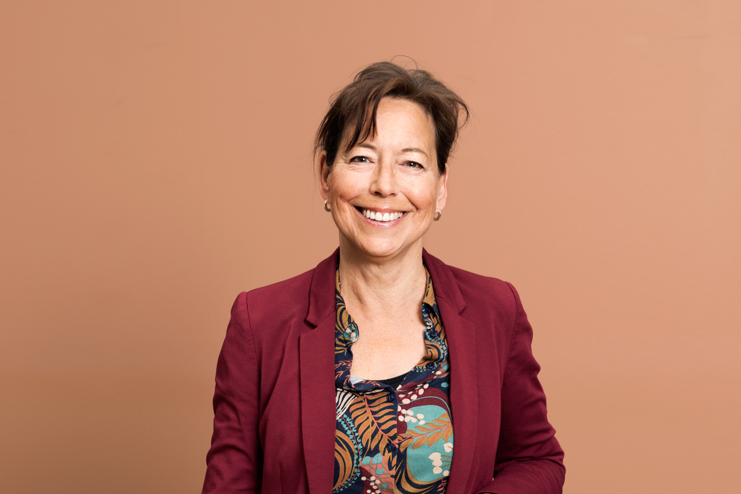 Anna-Karin Lindblom mot en terracottafärgad vägg. Anna-Karin har en röd kavaj och ser glad ut.