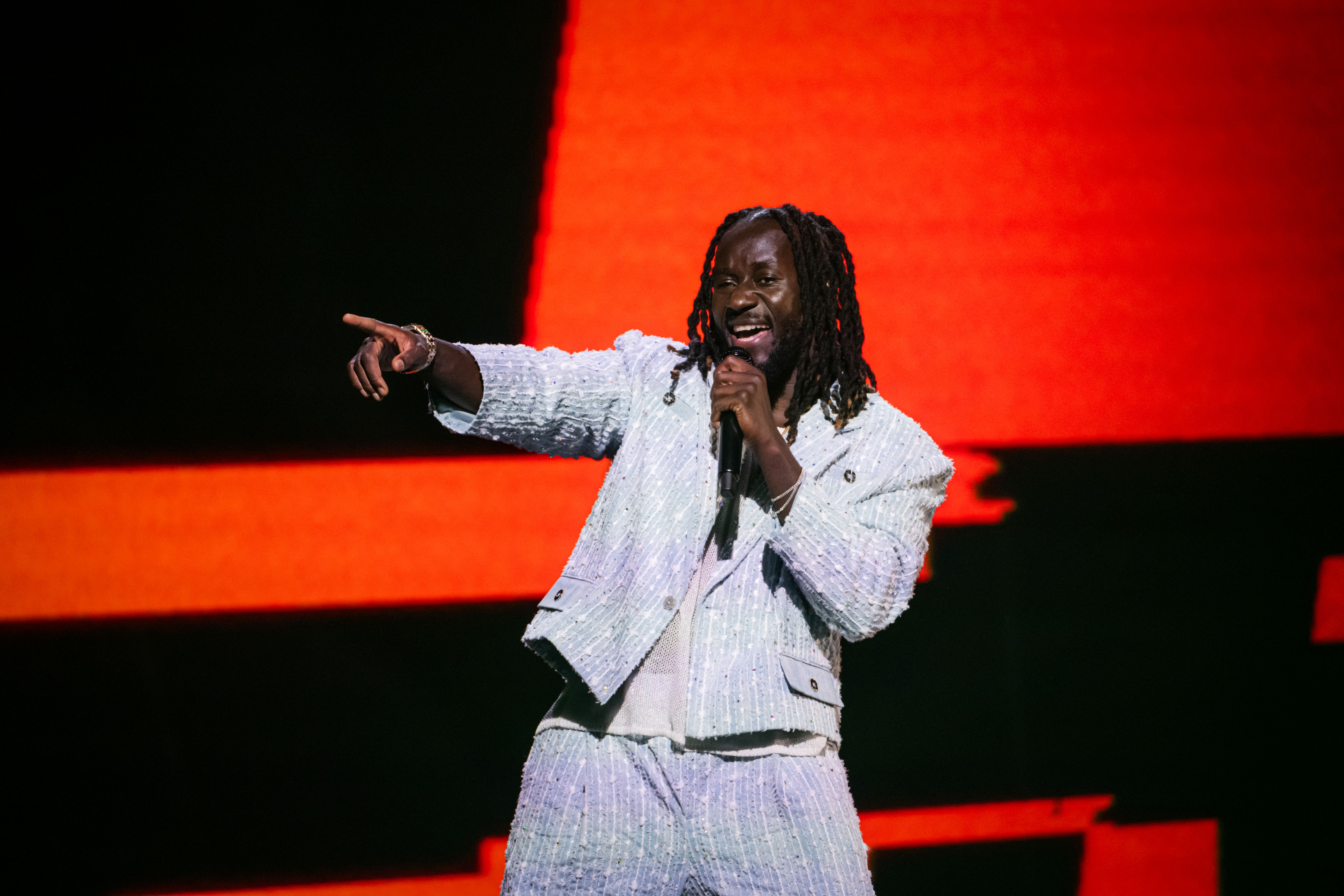 Pa Modou Badjie sjunger på scenen på Melodifestivalen. Han har ljusblå kostym och pekar mot någon ur bild.