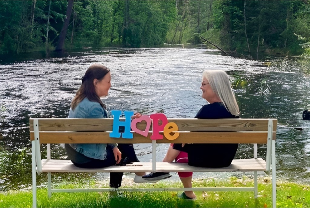 Två kvinnor sitter på en bänk vid vatten. På bänken står det "Hope" i färgglada bokstäver.