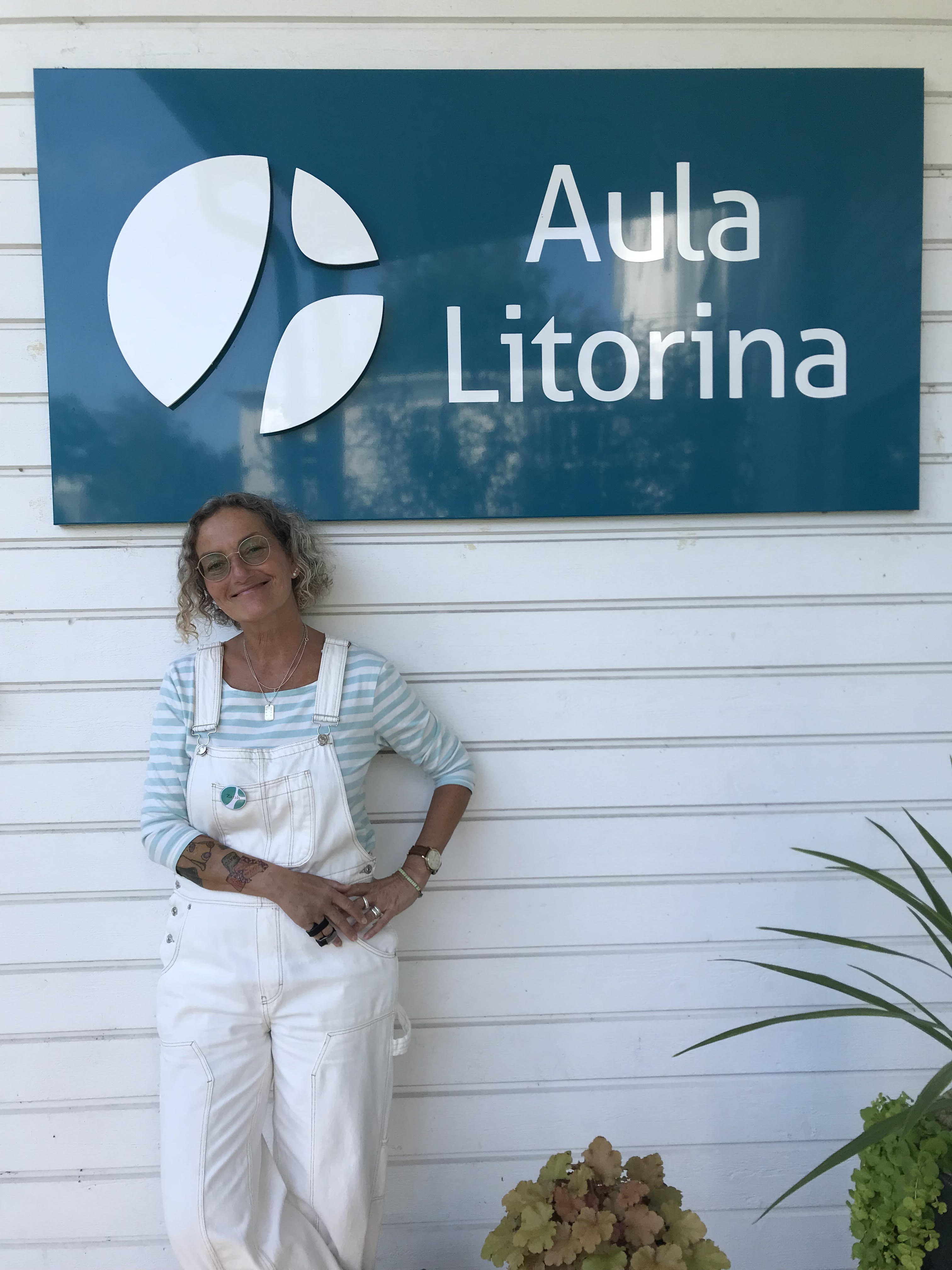 Läraren Zarah Necksten framför en vit vägg som har en stor turkos skylt där det står "Aula Litorina"