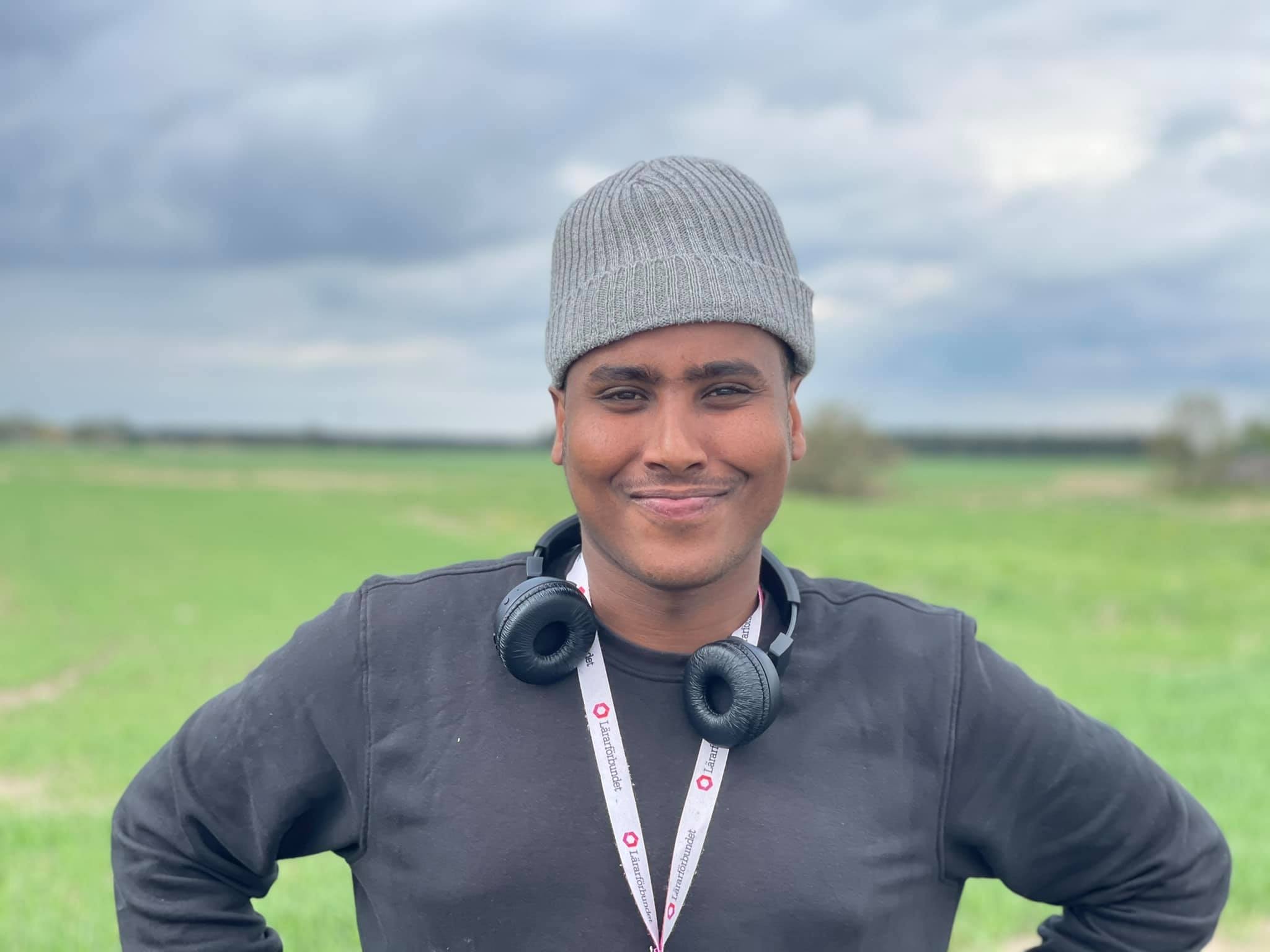 Faraah Mohamud står utomhus framför ett grönt fält och en blå lite molnig himmel. Han har ett par hörlurar kring halsen, ler och tittar rakt in i kameran.