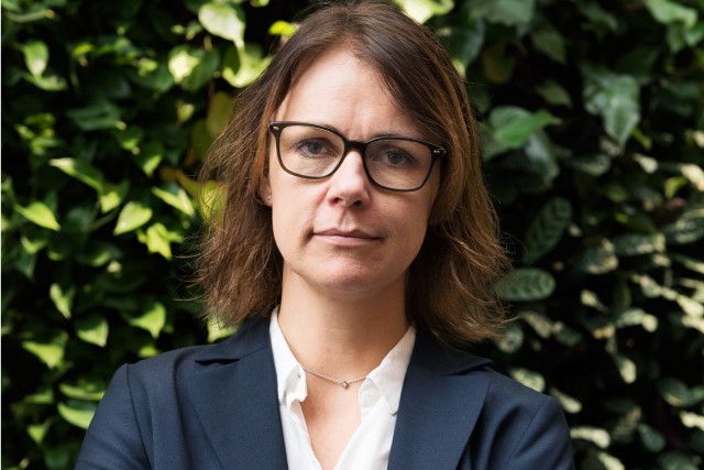 Pressbild Maria Graner. Maria ser allvarlig ut. Hon står framför en vägg full av växter.