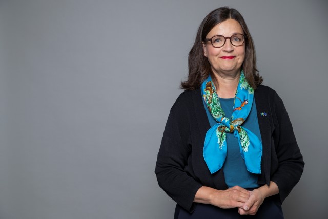 Pressbild Anna Ekström. Anna står till höger i bild framför en grå bakgrund. Hon har turkos blus och en mönstrad matchande knuten sjal runt halsen.