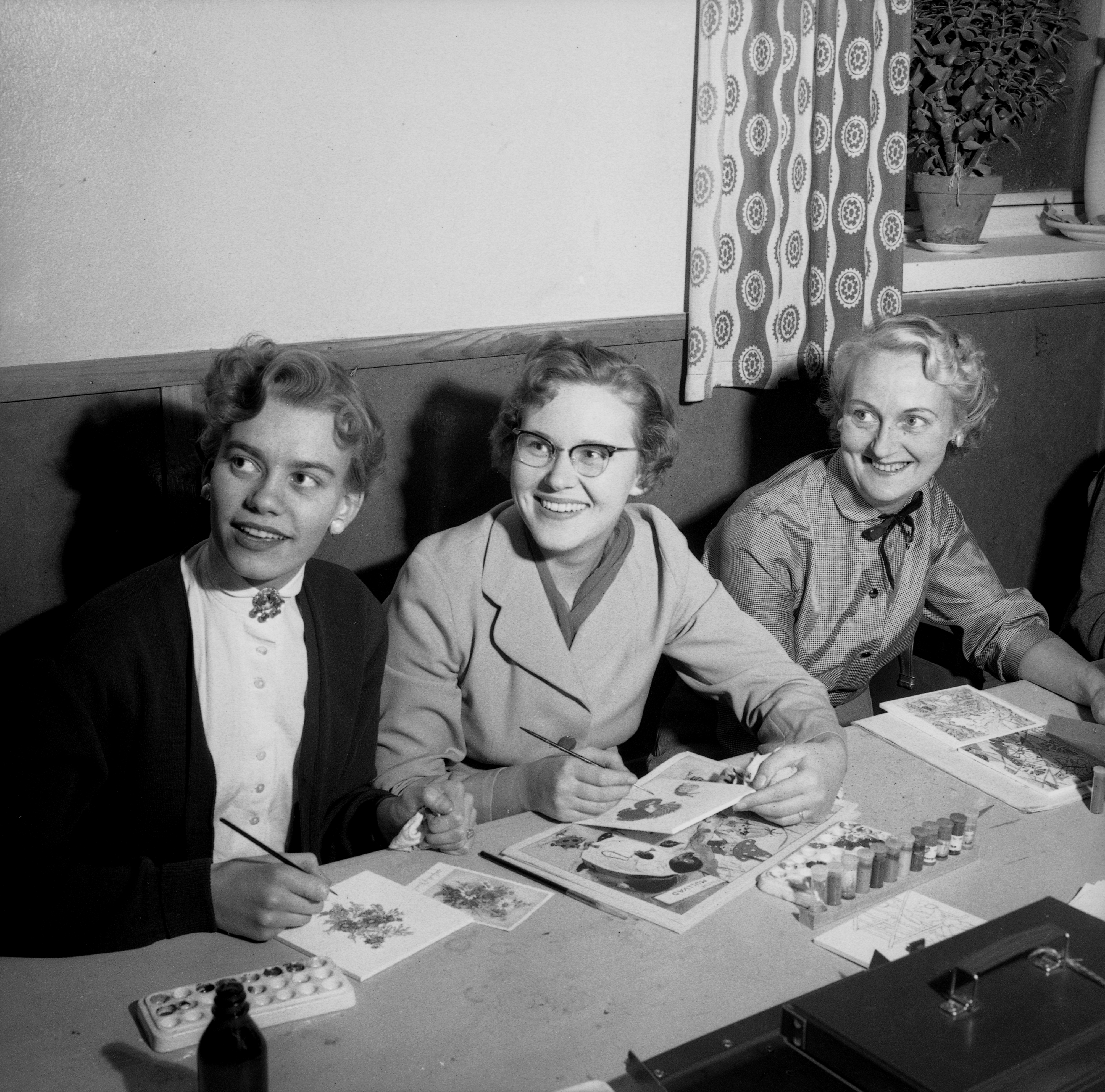 Svartvitt foto på tre personer som sitter vid ett bord och målar. De tittar upp åt sidan och ler.