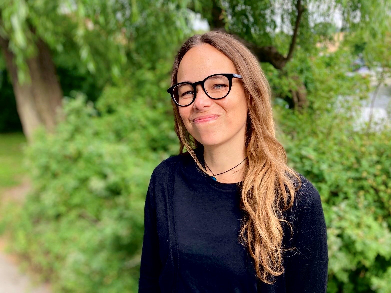 Porträttbild Jenny Holmberg. Jenny har långt vågigt brunt hår, glasögon och står utomhus med grönska i bakgrunden.