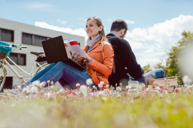 Två studenter som sitter på en gräsmatta och studerar. De sitter och lutar ryggarna mot varandra.
