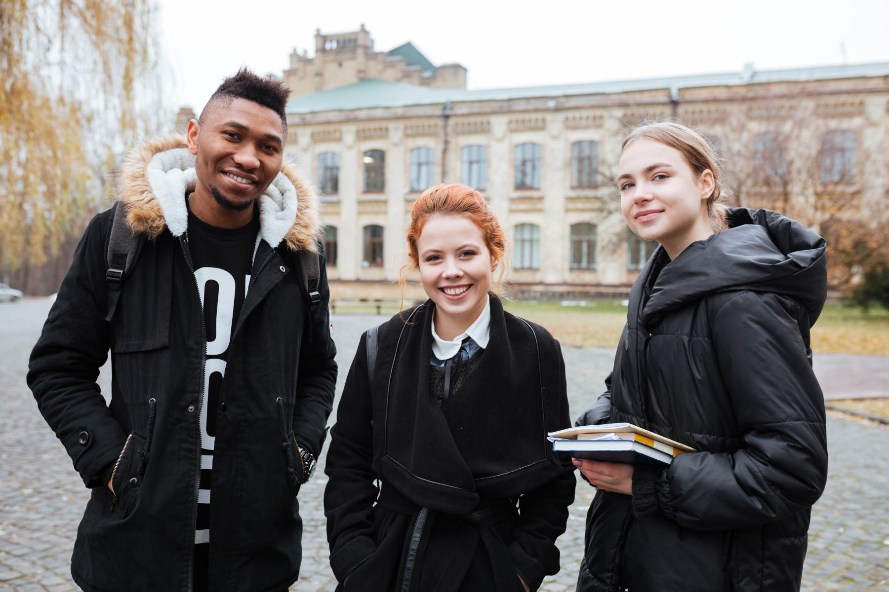 Tre unga studenter står framför en skolbyggnad, de ler mot kameran.