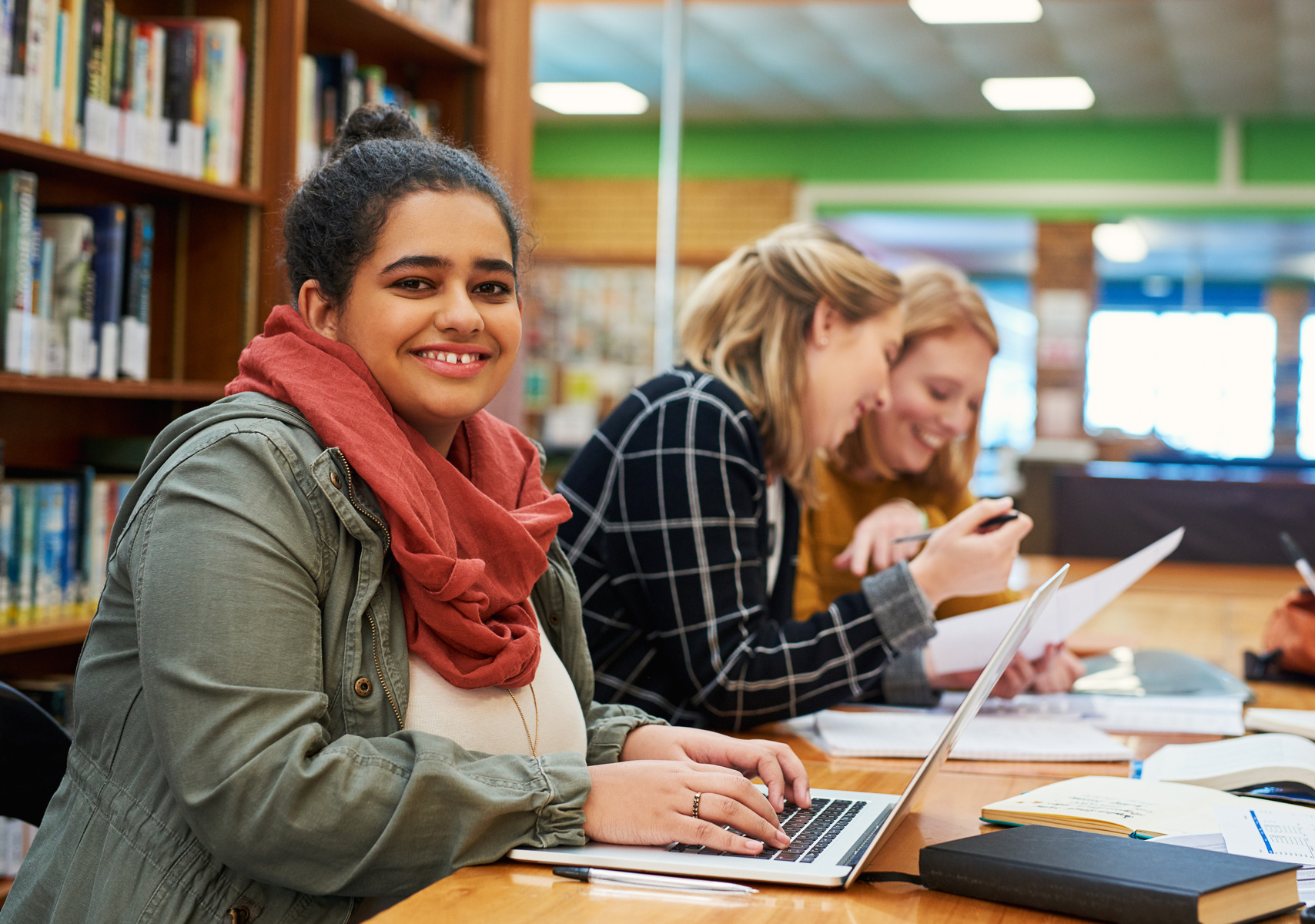 Tre personer sitter i ett bibliotek och studerar, två tittar ner i sina böcker och den tredje ser rakt in i kameran och ler.