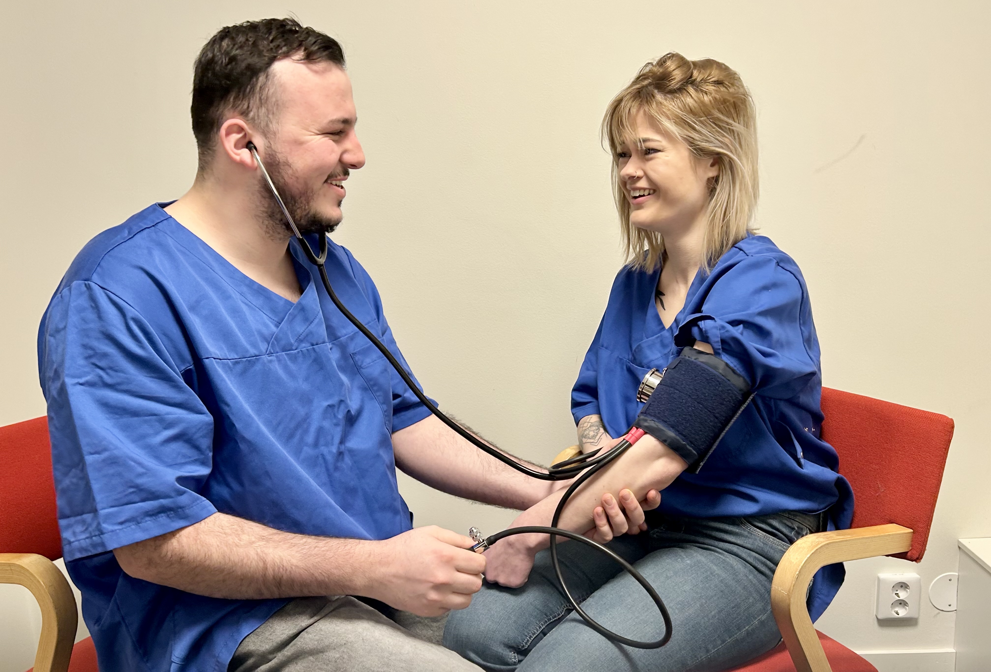 Youssef och en kvinna sitter iklädda blåa vårdkläder. Youssef tar blodtryck på kvinnan. De ler mot varandra.